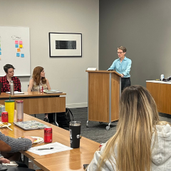A classroom shot shows a speaker at a podium addressing students