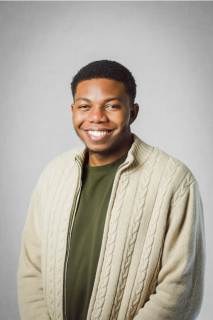 A studio portrait of Victor Olusegun shows him smiling into the camera against a grey backdrop, dressed in a dark green shirt with a beige cardigan over top