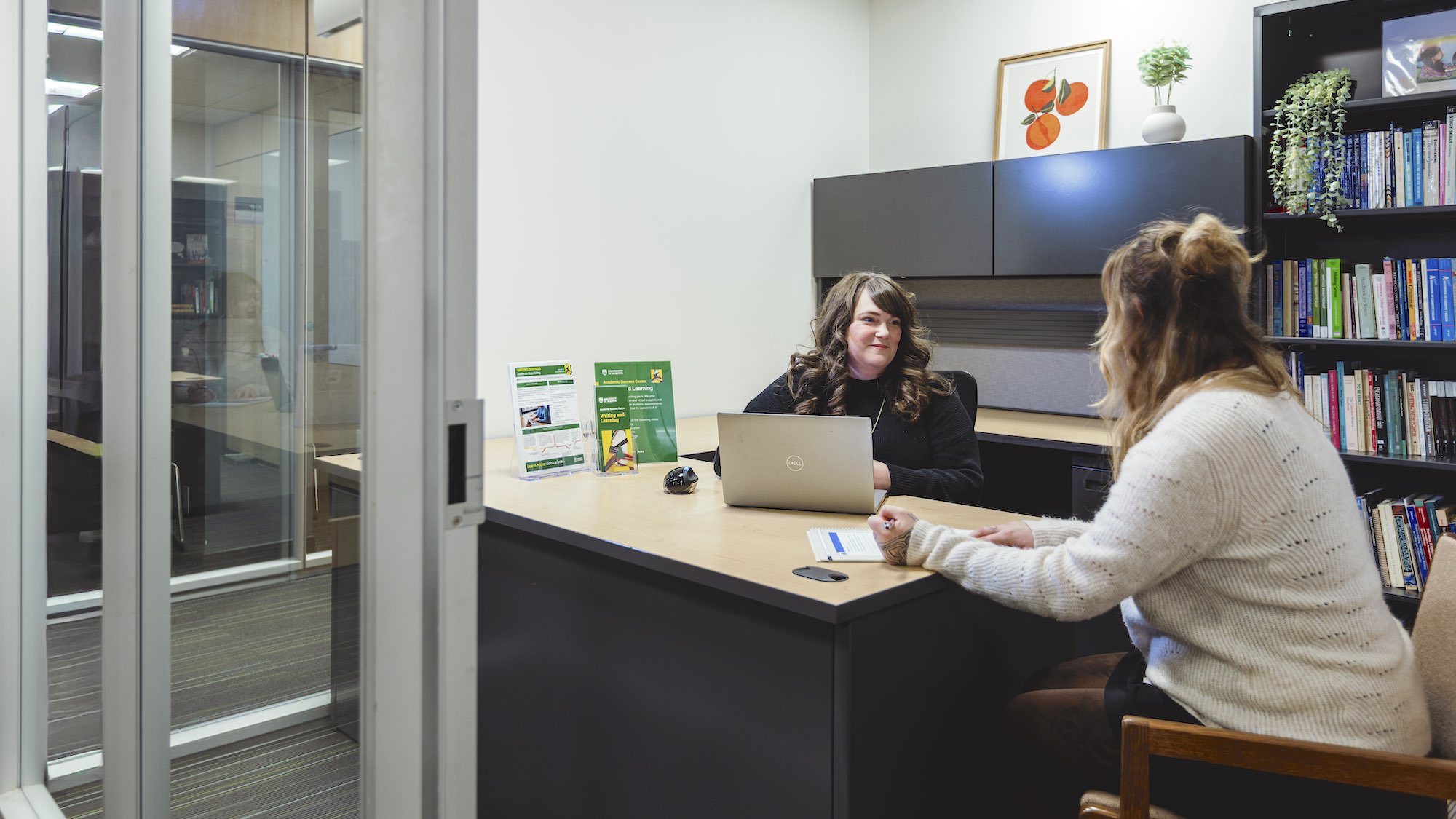 In an office, one woman behind a desk on a laptop listens to another woman there for an appointment with a pen in her hand ready to write