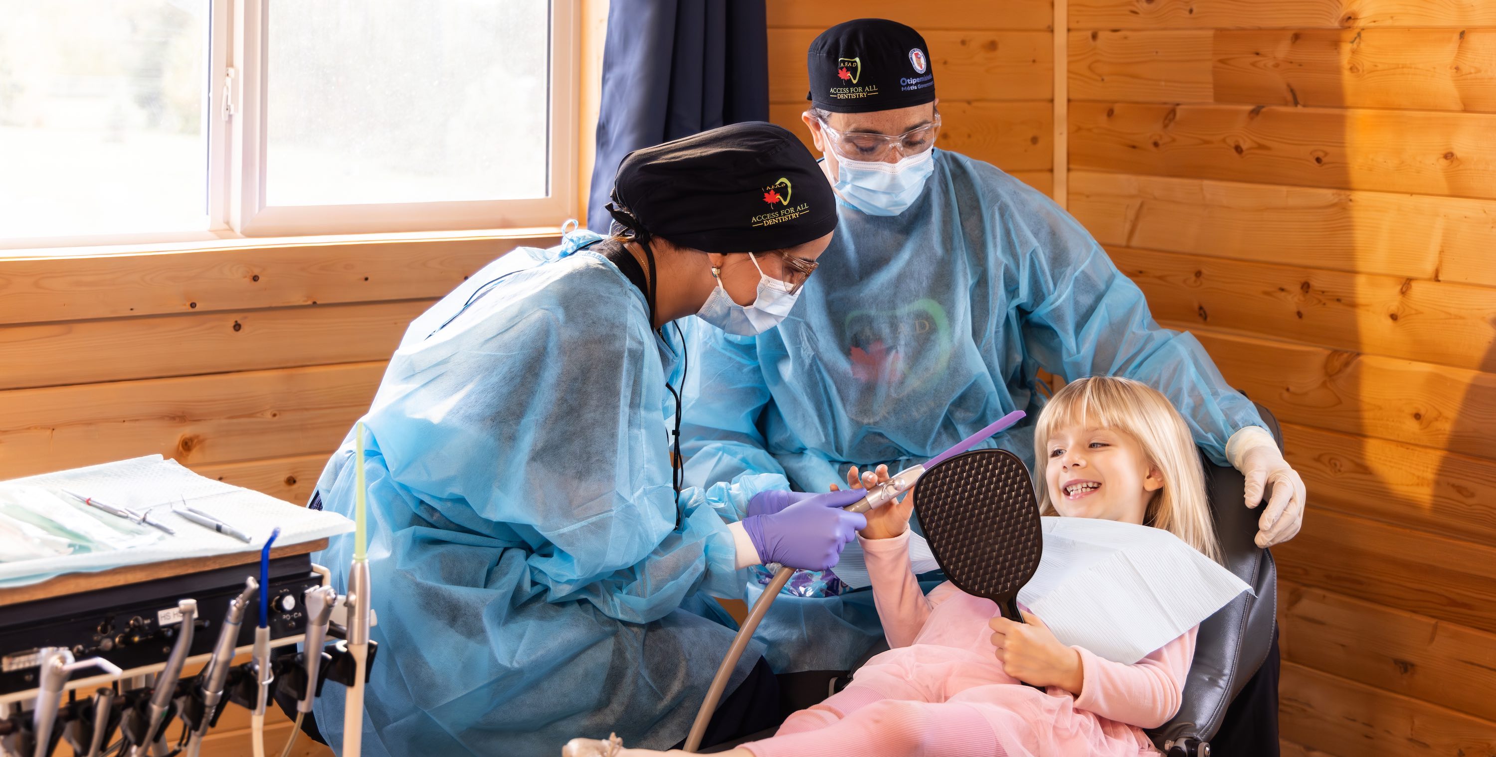A dental student supervised by a dentist holds a dental suction devide to a young girl on a dental chair who holds a mirror and looks at her teeth