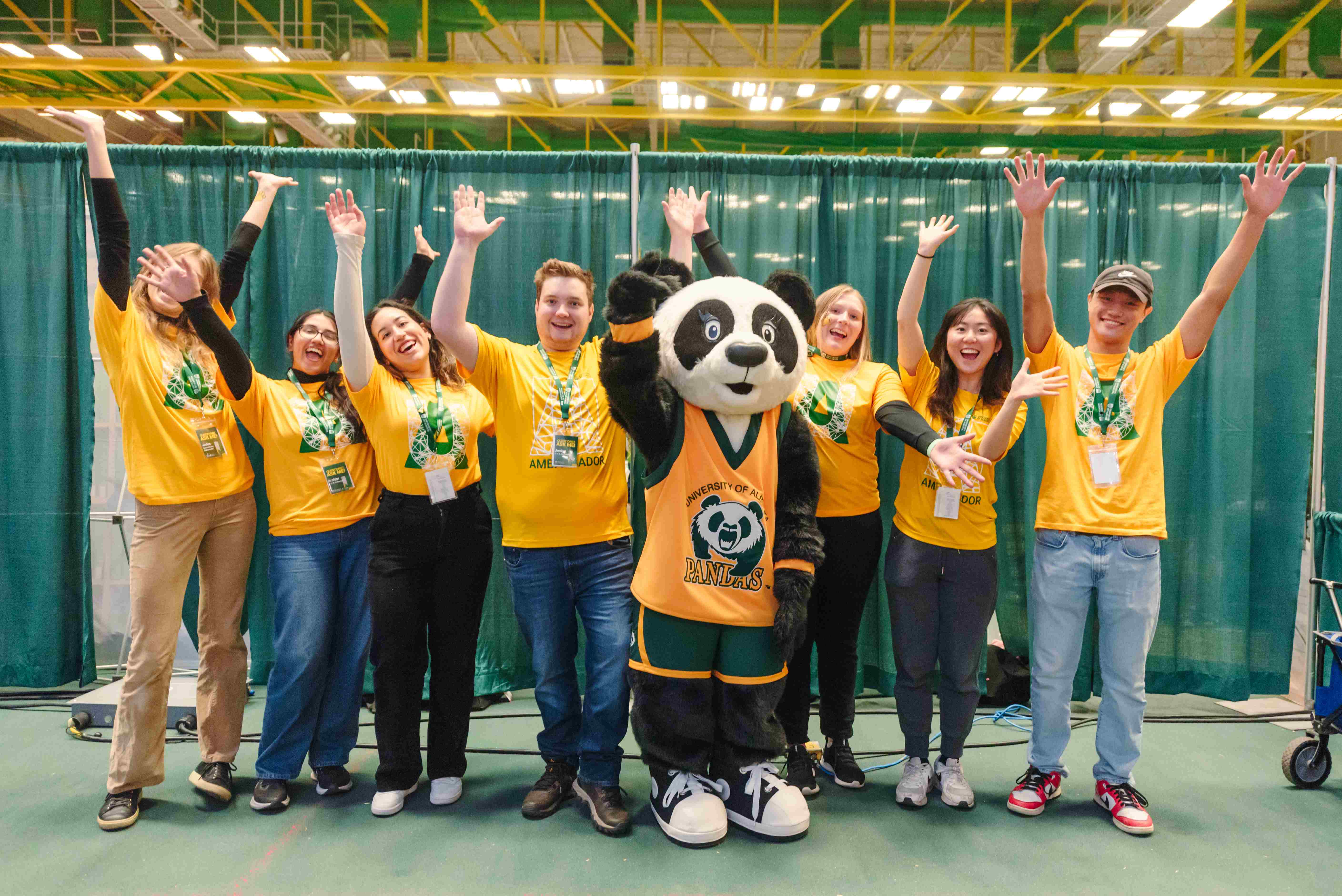 University of Alberta student ambassadors wearing yellow T-shirts pose enthusiastically with the university’s panda mascot at an indoor campus event.